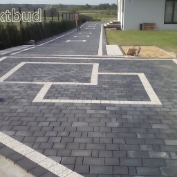Perfektbud Bartlomiej Nowakowski - Ułożona kostka brukowa w kolorze grafitowym z białymi elementami dekoracyjnymi, tworząca wzór geometryczny na podjeździe przed domem z widocznym trawnikiem i ogrodzeniem.