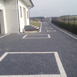 Perfektbud Bartlomiej Nowakowski - Nowoczesny podjazd z ciemnoszarej kostki brukowej z białymi obramowaniami, prowadzący do białego domu z garażem, w tle widoczny krajobraz z zielenią i ogrodzeniem.