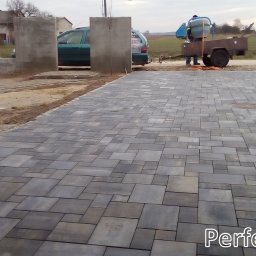 Perfektbud Bartlomiej Nowakowski - Ułożona kostka brukowa w odcieniach szarości na posesji, widoczna betoniarka na przyczepie i fragment budowy ogrodzenia.
