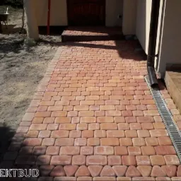 Ułożona ścieżka z czerwono-pomarańczowej kostki brukowej prowadząca do wejścia domu, z odpływem liniowym po prawej stronie i widocznym fragmentem trawnika po lewej.