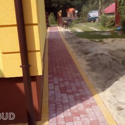 Perfektbud Bartlomiej Nowakowski - Świeżo ułożony chodnik z czerwonej kostki brukowej z żółtymi obrzeżami prowadzący wzdłuż żółtej ściany budynku, w tle widoczna taczka, betoniarka na przyczepce i samochód.