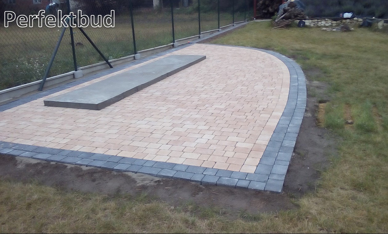 Wyłożony kostką brukową taras z szarą obwódką, widoczny betonowy element konstrukcyjny pośrodku, trawnik z tyłu, siatka ogrodzeniowa w tle.