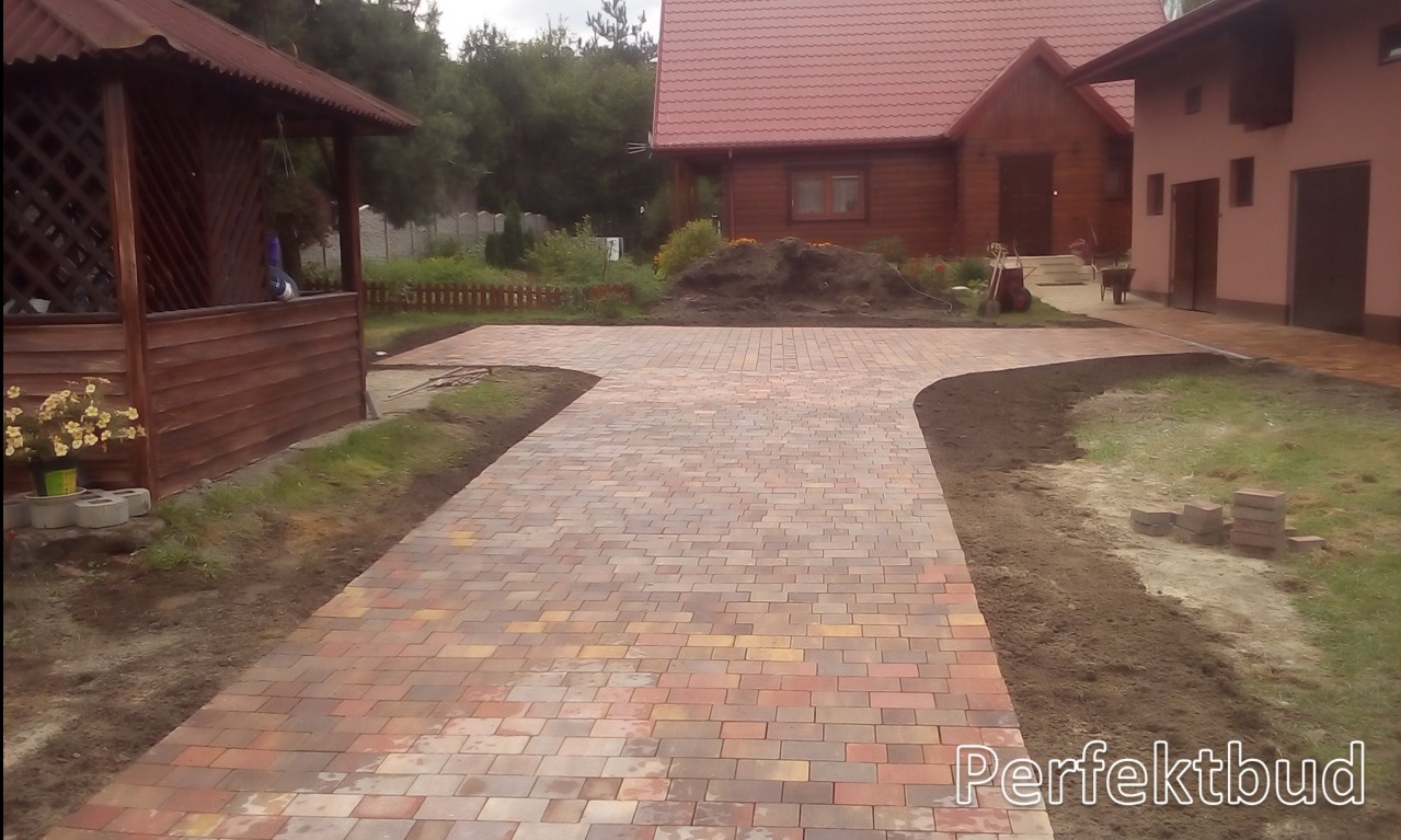 Nowo ułożona, wielokolorowa kostka brukowa tworząca ścieżkę i podjazd przed domem z drewnianą elewacją i altaną, z widocznymi stosami kostki brukowej i świeżo obsypaną ziemią po bokach.