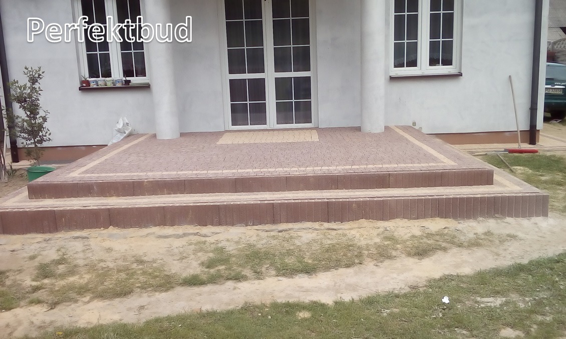 Schody wejściowe do domu wyłożone brązową kostką brukową z jasną obwódką, widok na białą fasadę budynku z oknami i kolumnami.