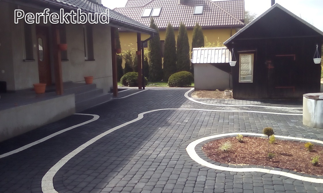 Czarno-szara kostka brukowa ułożona w serpentynowy wzór z białymi obramowaniami, prowadząca do domu z werandą i małej, drewnianej budowli z doniczkami, na tle zielonych tuj.