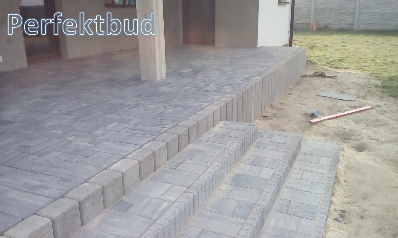 Szara kostka brukowa ułożona na tarasie i schodach, widoczne krawężniki i nierówności terenu obok, poziomica leżąca na piasku.