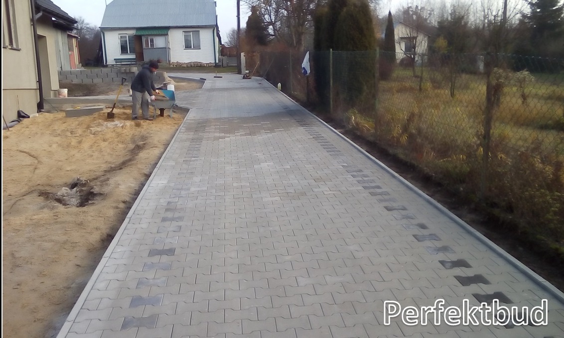 Układanie szarej kostki brukowej typu I na posesji, widoczny pracownik przy betoniarce i częściowo ułożona nawierzchnia z ciemniejszymi elementami dekoracyjnymi po bokach.
