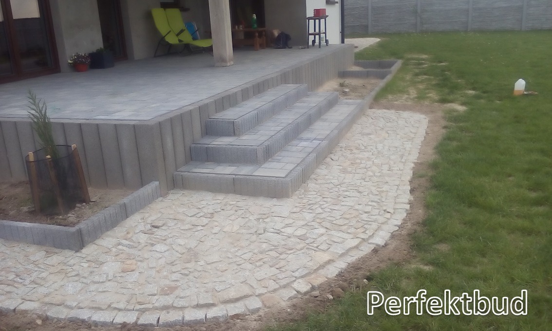 Wykończony taras z kostki brukowej z kamiennymi schodami i ścieżką z kostki granitowej, obrzeża z betonowych krawężników, widok na trawnik i dom z zielonymi krzesłami na tarasie.