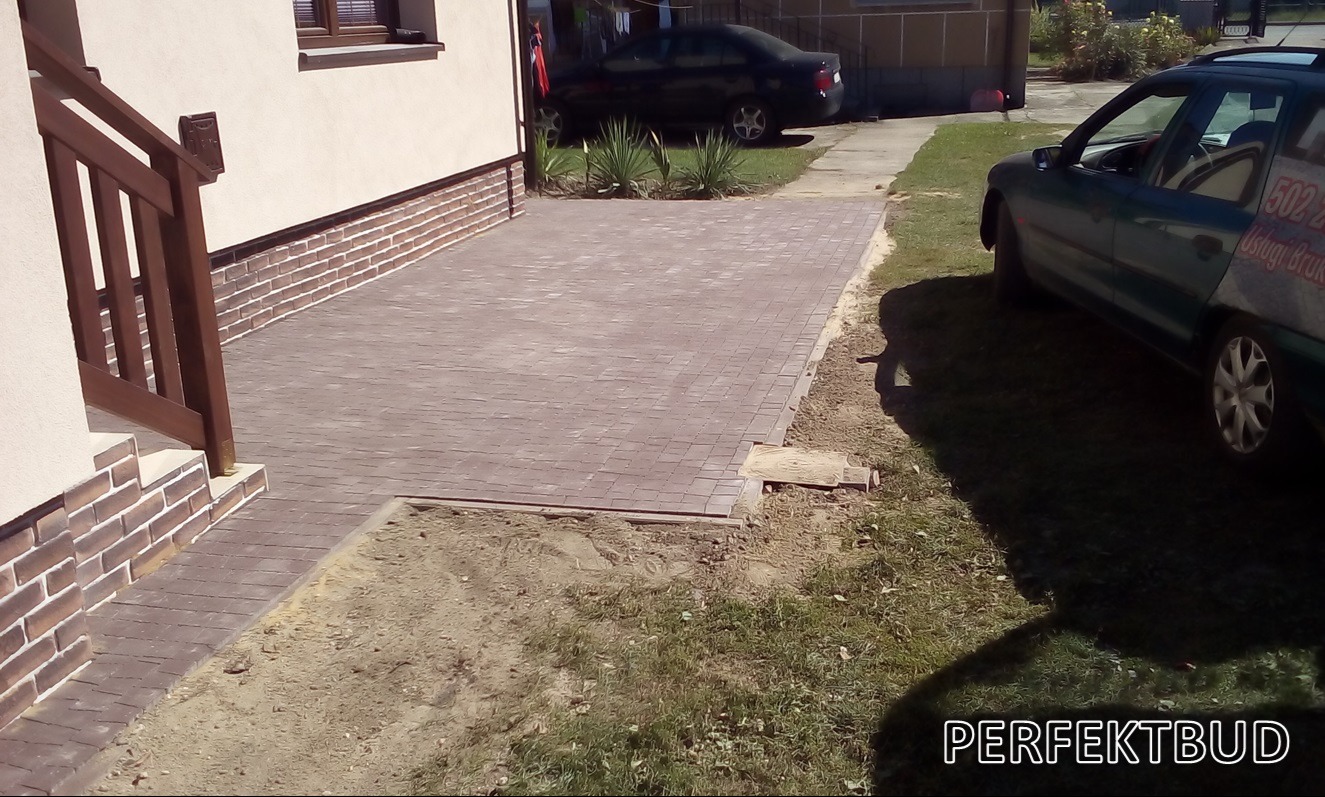 Ułożona, brązowa kostka brukowa przed domem z brązową podmurówką i drewnianą balustradą schodów wejściowych. Widoczny fragment trawnika i zaparkowane samochody.