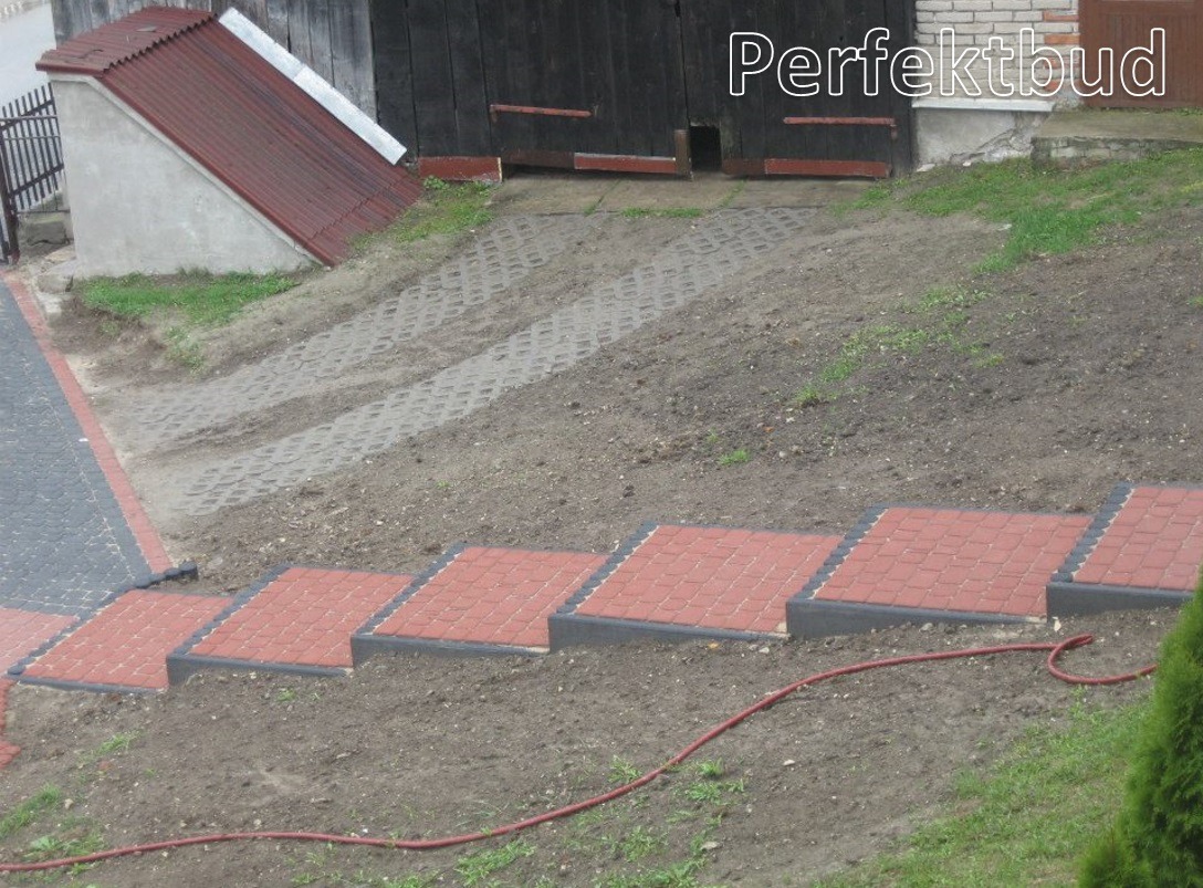 Schody z czerwonej kostki brukowej z czarnym obramowaniem prowadzące w górę zbocza, w tle widoczna brama garażowa i betonowy schron z metalowym dachem.