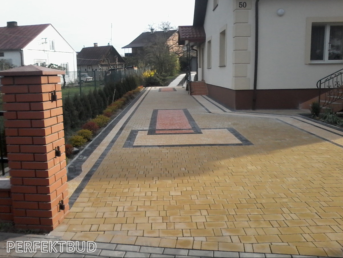Żółta kostka brukowa ułożona w geometryczny wzór na podjeździe przed domem z numerem 50, obramowana czarną kostką i ceglanym słupkiem.
