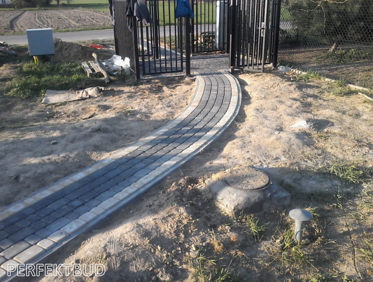 Zakrzywiona ścieżka z szarej kostki brukowej prowadząca do czarnej metalowej bramy, widoczna studzienka kanalizacyjna po prawej stronie, teren jeszcze niezagospodarowany.