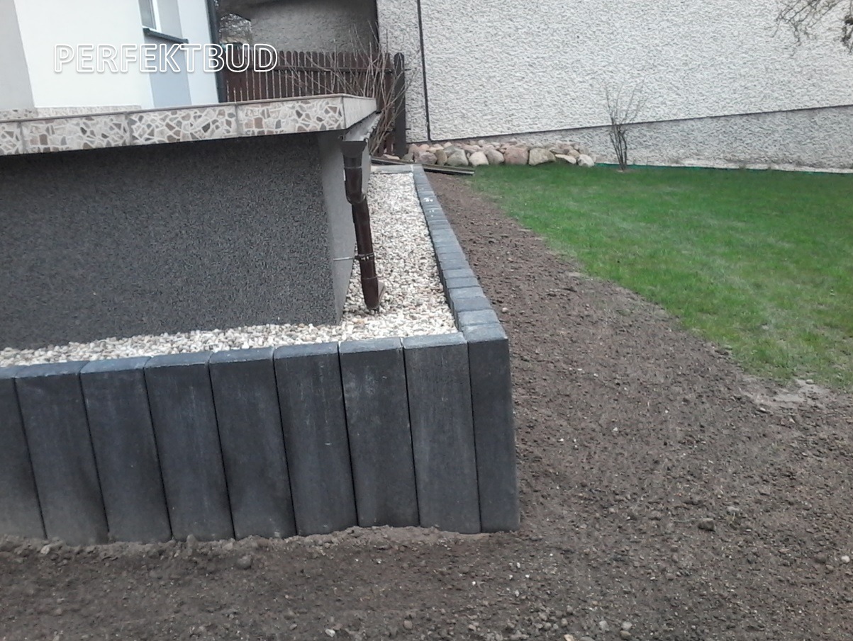 Szara kostka brukowa obrzeżająca rabatę z białym żwirem, widoczny fragment trawnika i elewacji budynku.