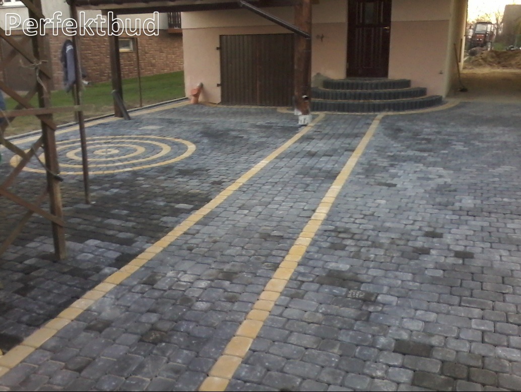 Ułożona kostka brukowa w kolorze szarym z żółtymi akcentami tworzącymi wzór okręgu i linii, prowadząca do budynku z bramą garażową i schodami, widoczny częściowo drewniany szkielet z lewej strony.