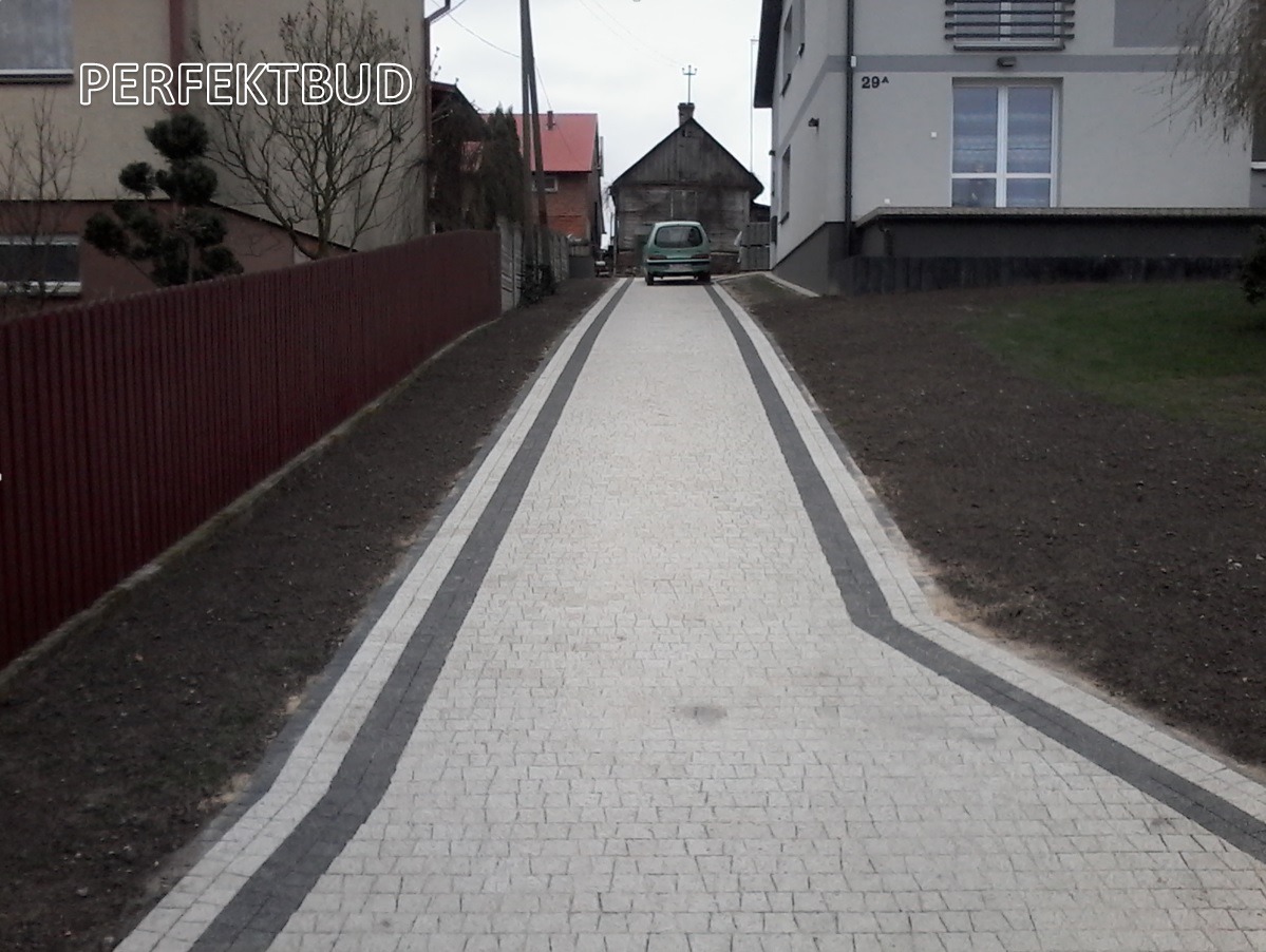 Długa, lekko zakrzywiona ścieżka z jasnoszarej kostki brukowej z ciemnoszarymi obrzeżami, prowadząca do domu z numerem 29A i zaparkowanego samochodu na końcu posesji.