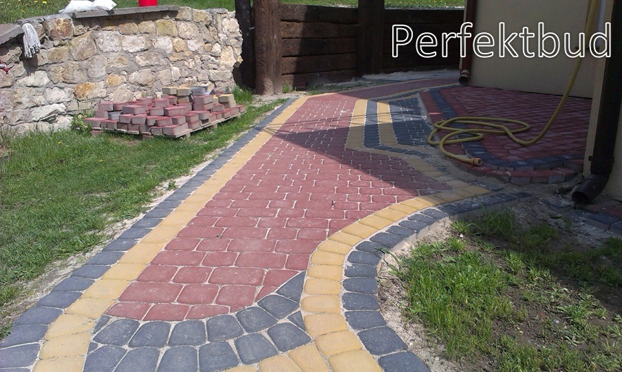 Ścieżka z kostki brukowej w odcieniach czerwieni, żółci i grafitu, z paletą kostki przygotowanej do układania przy kamiennym murku w tle.