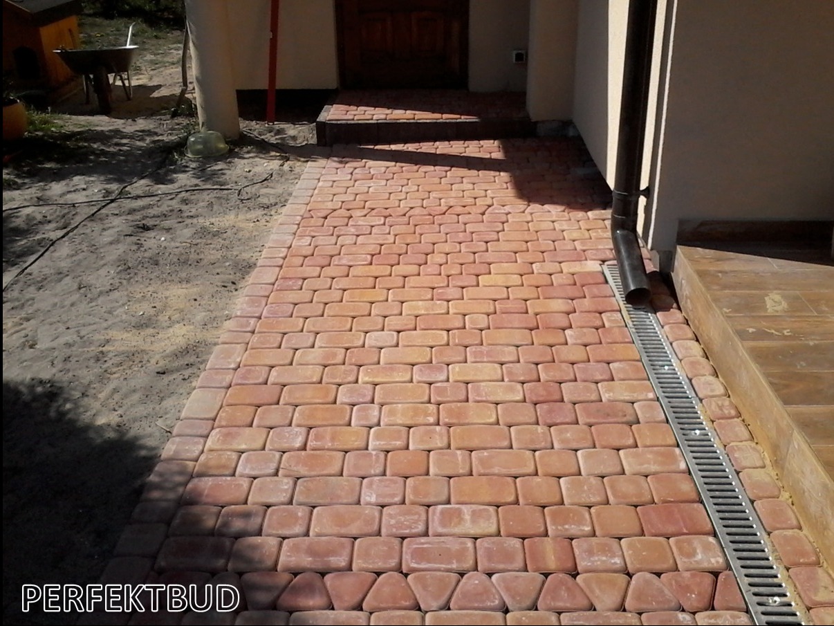 Ułożona ścieżka z czerwono-pomarańczowej kostki brukowej prowadząca do wejścia domu, z odpływem liniowym po prawej stronie i widocznym fragmentem trawnika po lewej.