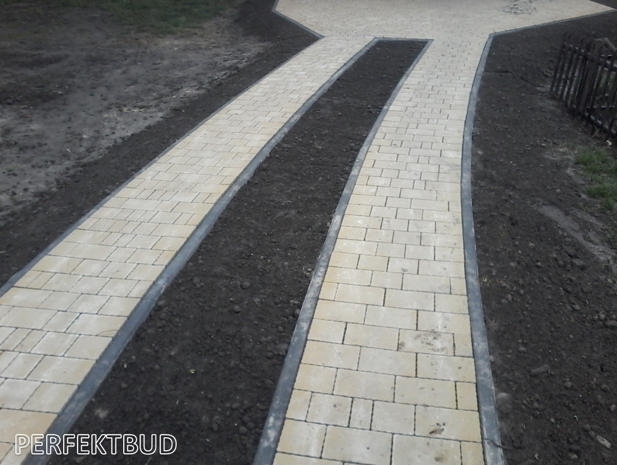 Rozchodzące się ścieżki z jasnej kostki brukowej, oddzielone pasem czarnej ziemi, z widocznym ogrodzeniem w tle.