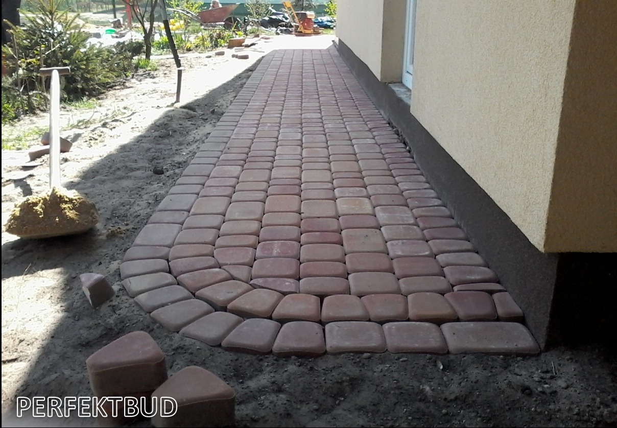 Świeżo ułożona ścieżka z czerwonej kostki brukowej prowadząca wzdłuż ściany budynku, widoczna łopata z piaskiem i kilka nieułożonych kostek.