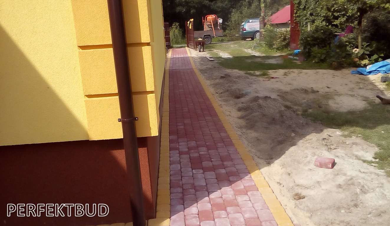 Świeżo ułożony chodnik z czerwonej kostki brukowej z żółtymi obrzeżami prowadzący wzdłuż żółtej ściany budynku, w tle widoczna taczka, betoniarka na przyczepce i samochód.