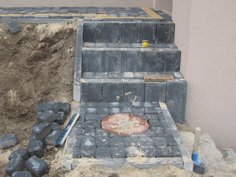 Budowa schodów z kostki brukowej w kolorze grafitowym, z elementem dekoracyjnym w formie okrągłej rozety z czerwonej kostki, widoczne narzędzia i niedokończone prace ziemne.