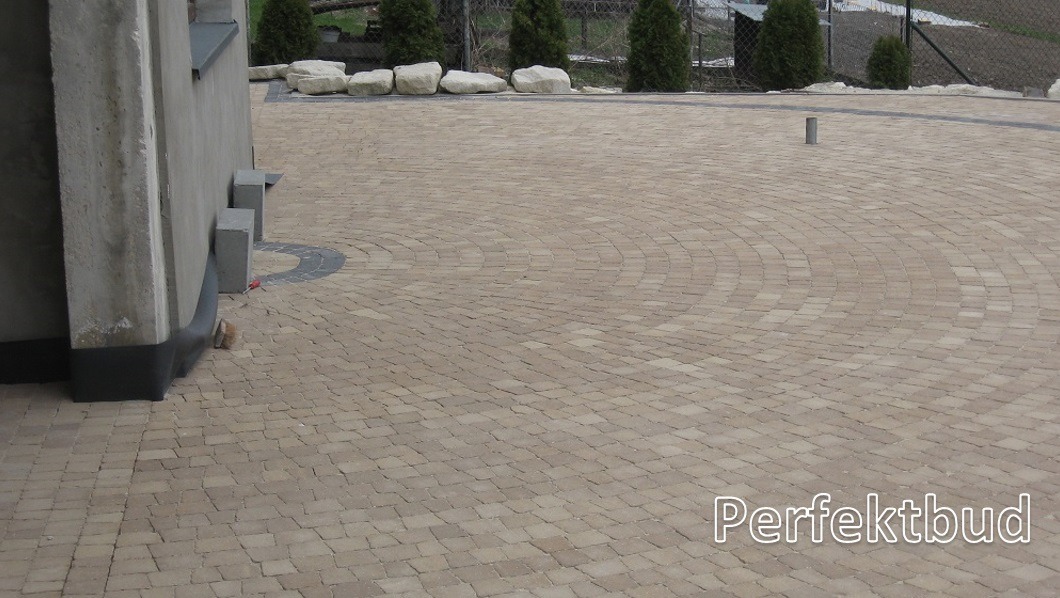 Beżowa kostka brukowa ułożona w półokręgi na podjeździe, widoczne elementy domu i ozdobne kamienie w tle.