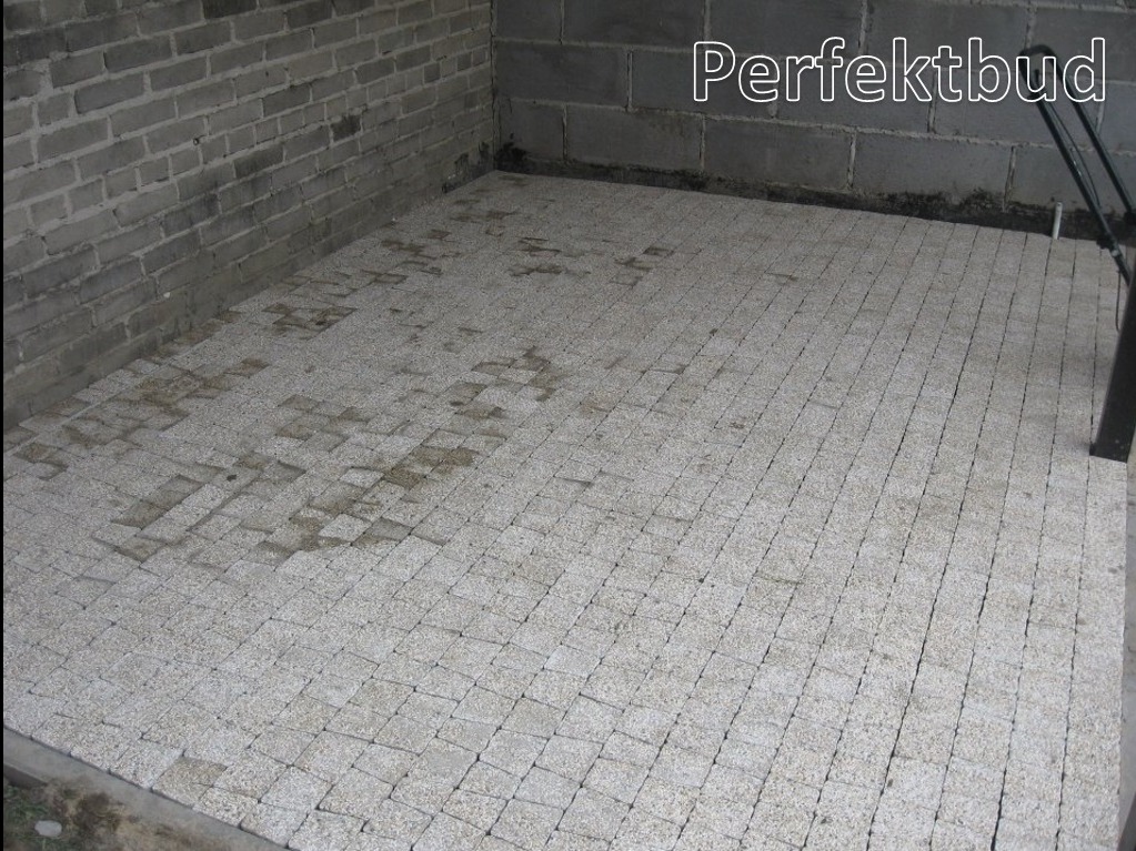 Nowo ułożona, jasnoszara kostka brukowa w pomieszczeniu w Kazimierzu Dolnym, z widocznymi śladami wilgoci i fragmentami ściany z szarej cegły. Widoczny cień stojaka w prawym górnym rogu.