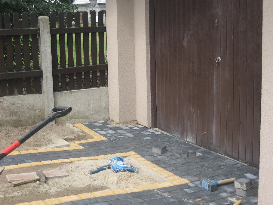 Częściowo ułożona kostka brukowa w kolorze szarym z żółtą obwódką przed brązową bramą garażową, widoczne narzędzia: młotek brukarski, łopata i poziomica.