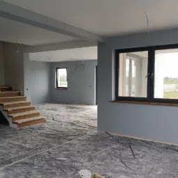 Wnętrze domu w Jaworznie w trakcie prac wykończeniowych; ściany pomalowane na szaro, podłoga zabezpieczona folią, widoczne puszki z farbą i fragment schodów.