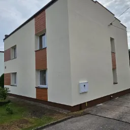 Otynkowana elewacja budynku z elementami dekoracyjnymi imitującymi drewno wokół okien, widoczna skrzynka elektryczna na ścianie.