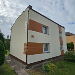 Dwupiętrowy budynek po remoncie elewacji z dekoracyjnymi poziomymi pasami imitującymi drewno i numerem domu 35, otoczony zielenią.