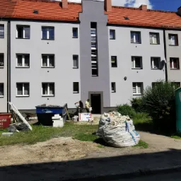 Pracownicy budowlani malują szarą elewację bloku mieszkalnego z czerwoną dachówką, widoczne worki z gruzem i kontener na śmieci.