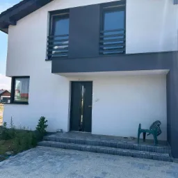 Nowoczesny dom jednorodzinny z białą elewacją, grafitowymi akcentami, balustradami okiennymi z poziomych profili, schodami wejściowymi wyłożonymi kostką brukową i zielonym krzesłem ogrodowym przed...