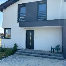 Nowoczesny dom jednorodzinny z białymi ścianami, grafitowymi elementami wykończeniowymi, szarą kostką brukową przed wejściem i zielonym krzesłem ogrodowym.