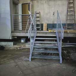 Limt Manufacturing - Metalowe schody z kratownicowymi stopniami i poręczami na tle surowego wnętrza hali w trakcie budowy lub remontu, z widocznymi drabinami, puszką farby i narzędziami.