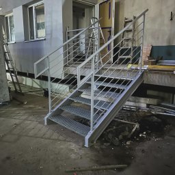 Limt Manufacturing - Metalowe schody z ażurowymi stopniami i prostą balustradą na tle szarego budynku i otwartych drzwi, widoczne drabiny i elementy konstrukcyjne.