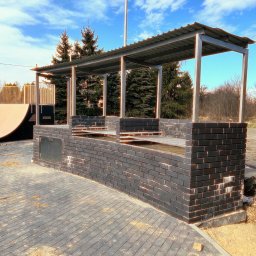 Limt Manufacturing - Murowana, zadaszona konstrukcja grilla z czarnej cegły z metalowymi elementami i szafką, na wybrukowanym patio, z rampą i drzewami w tle, pod błękitnym niebem.