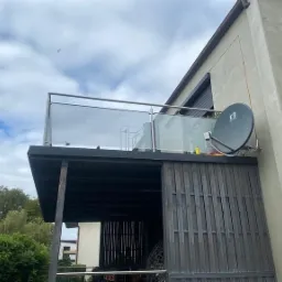 Nowoczesny balkon z balustradą ze stali nierdzewnej i szkła, widok z dołu na szarą elewację domu z anteną satelitarną i drewnianą konstrukcją pod spodem.