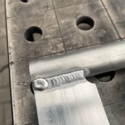 Detal spoiny łączącej aluminiowy profil okrągły z płaskownikiem na perforowanym stole spawalniczym.