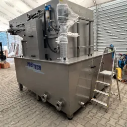 Szara przemysłowa maszyna z napisem POSITECH, częściowo owinięta folią, stoi na kostce brukowej obok aluminiowej drabiny, w tle widoczna hala namiotowa i inne urządzenia.