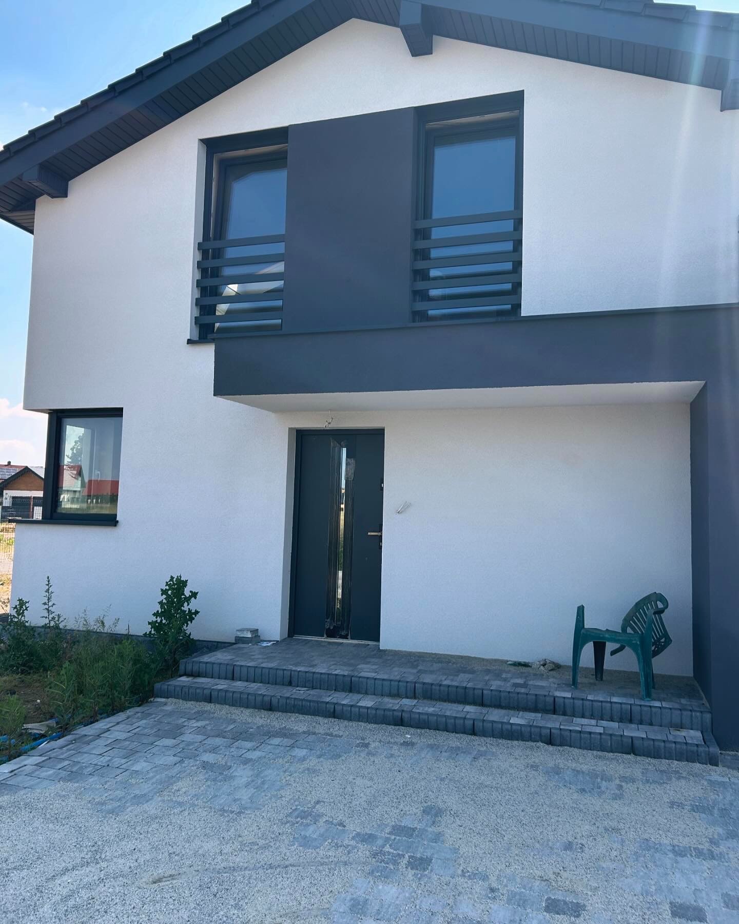 Nowoczesny dom jednorodzinny z białą elewacją, grafitowymi akcentami, balustradami okiennymi z poziomych profili, schodami wejściowymi wyłożonymi kostką brukową i zielonym krzesłem ogrodowym przed...