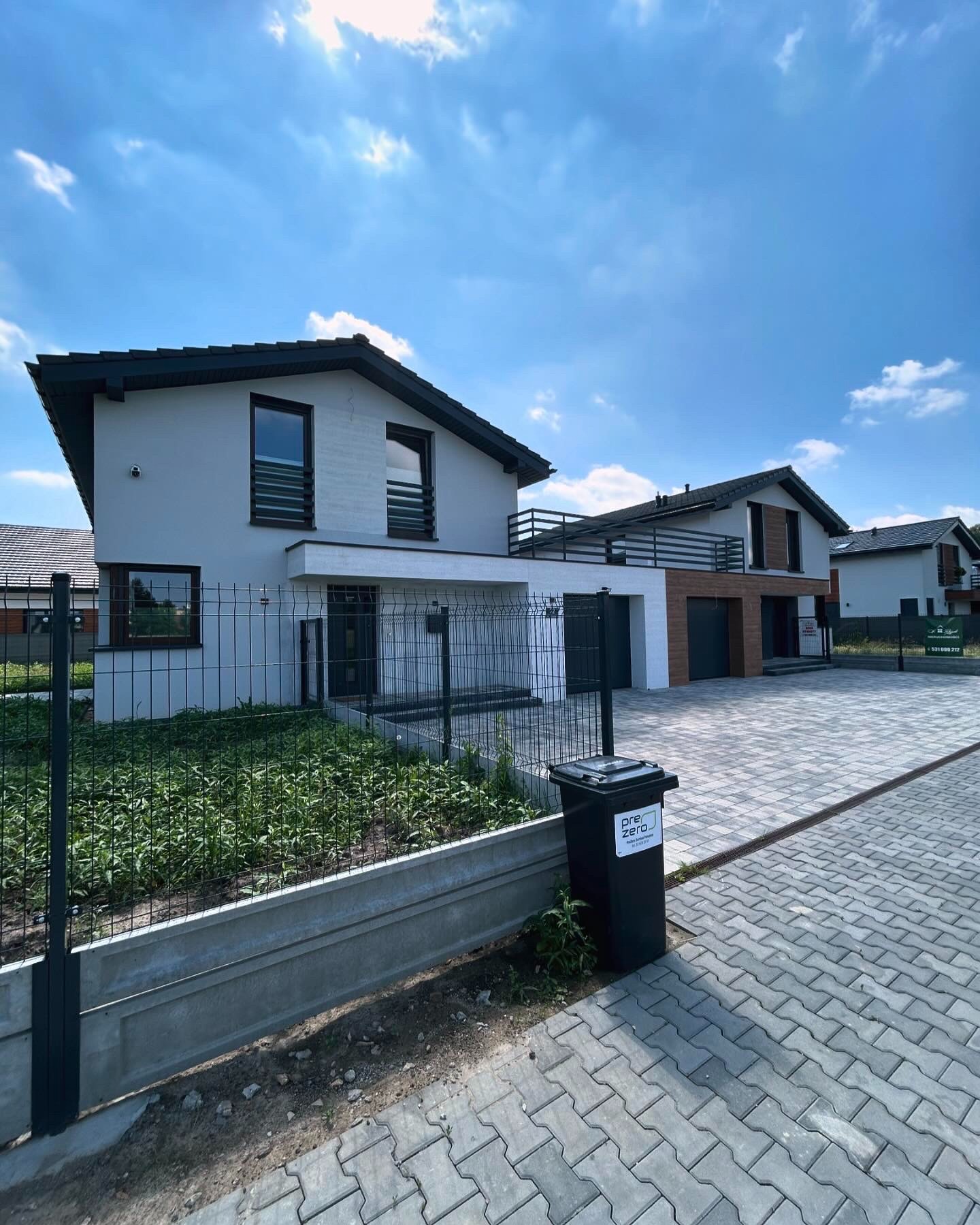 Nowoczesny dom jednorodzinny z szarą elewacją i czarnymi detalami, widoczny z zewnątrz przez metalowe ogrodzenie z podmurówką, na pierwszym planie czarny kosz na śmieci z naklejką.