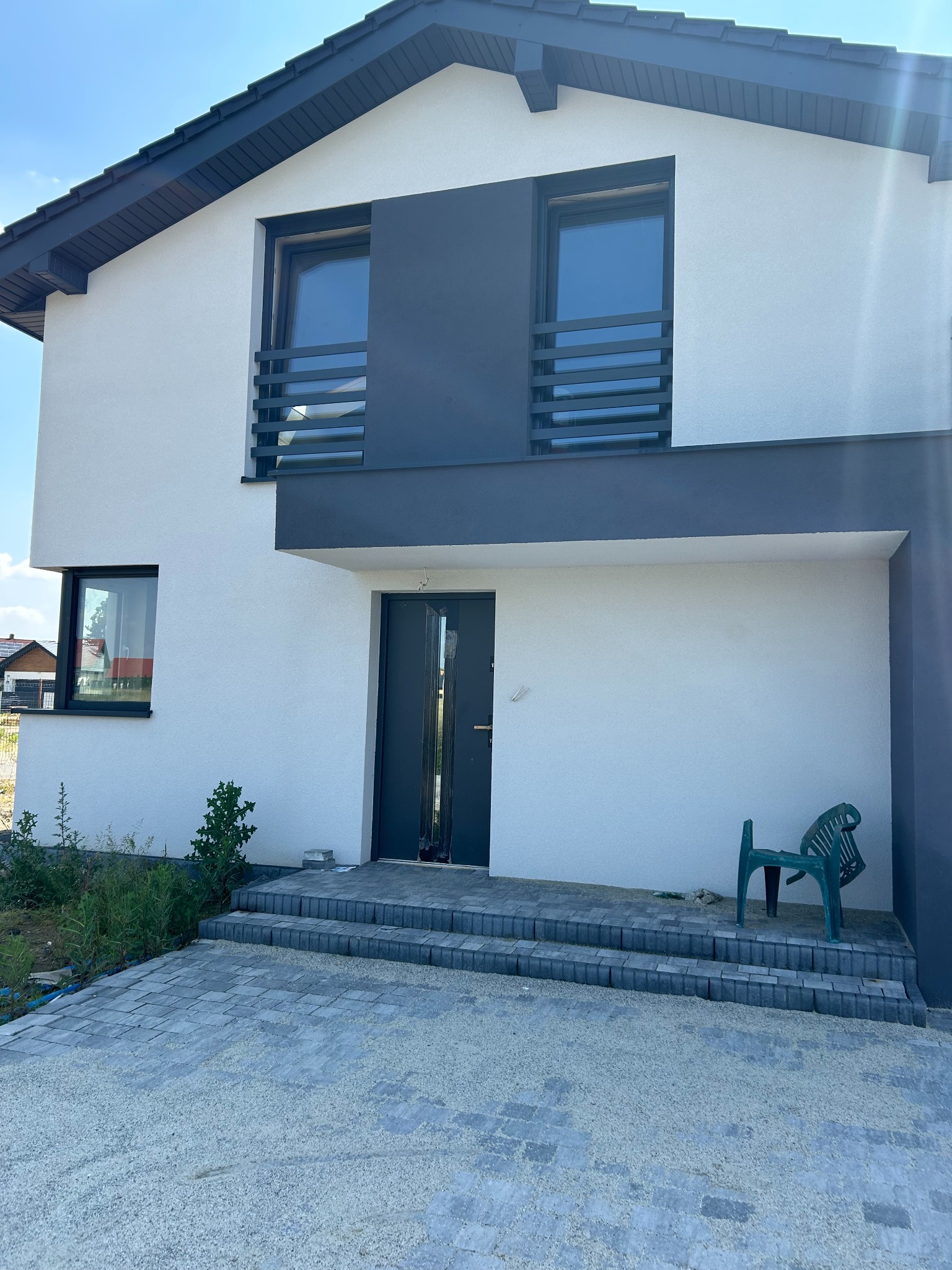 Nowoczesny dom jednorodzinny z białymi ścianami, grafitowymi elementami wykończeniowymi, szarą kostką brukową przed wejściem i zielonym krzesłem ogrodowym.