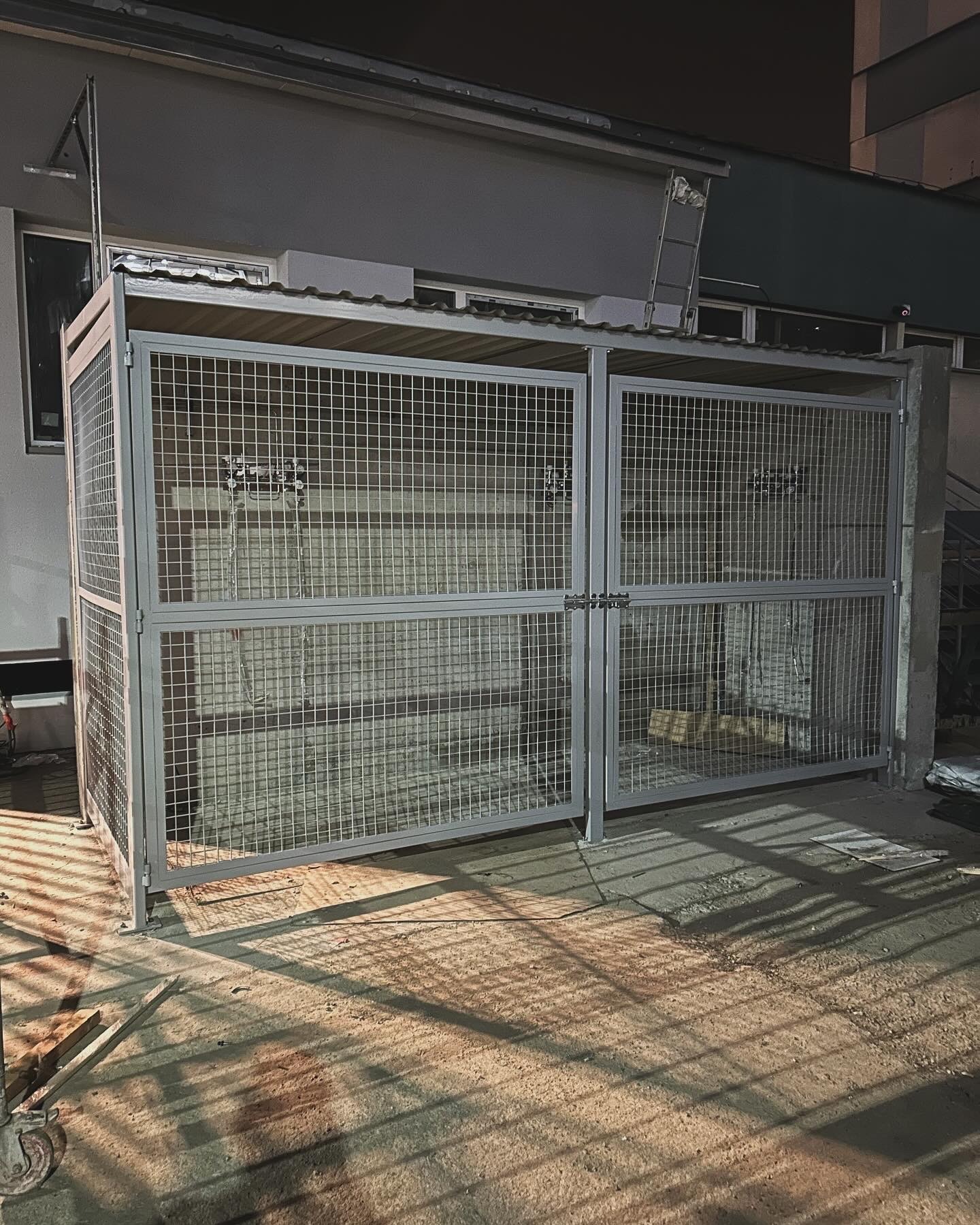 Metalowa klatka zamykana na kłódki, prawdopodobnie na śmieci, z widoczną konstrukcją nośną i siatką, umieszczona na zewnątrz budynku.