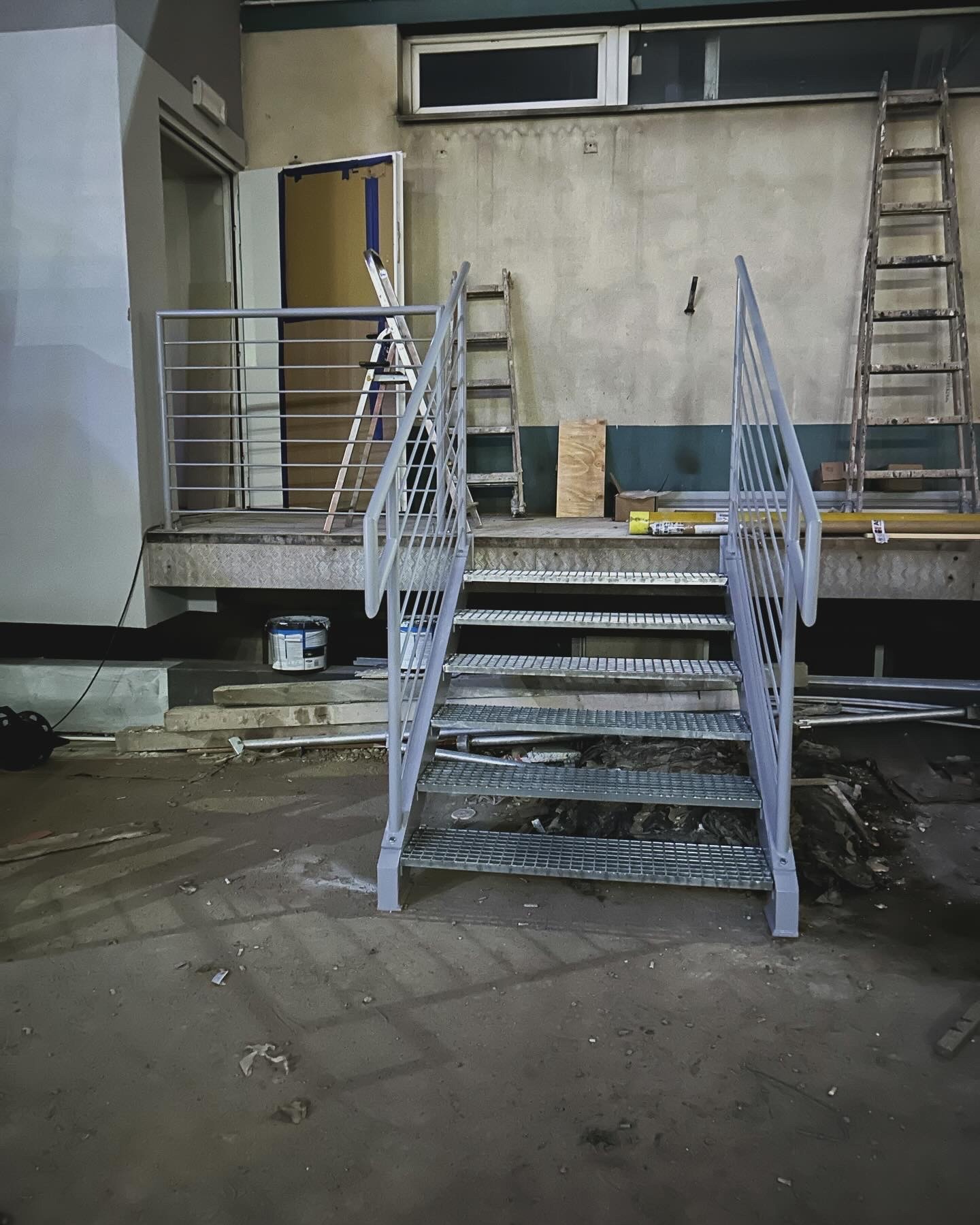 Metalowe schody z kratownicowymi stopniami i poręczami na tle surowego wnętrza hali w trakcie budowy lub remontu, z widocznymi drabinami, puszką farby i narzędziami.