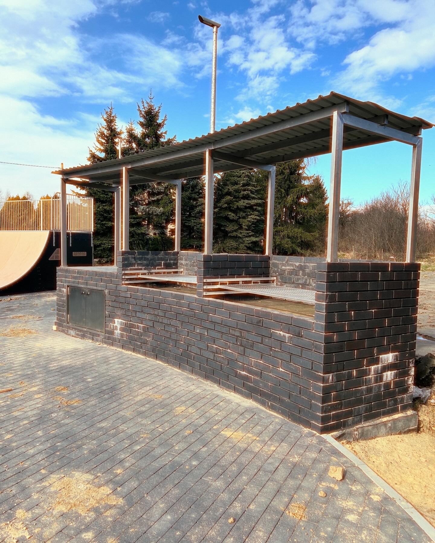 Murowana, zadaszona konstrukcja grilla z czarnej cegły z metalowymi elementami i szafką, na wybrukowanym patio, z rampą i drzewami w tle, pod błękitnym niebem.