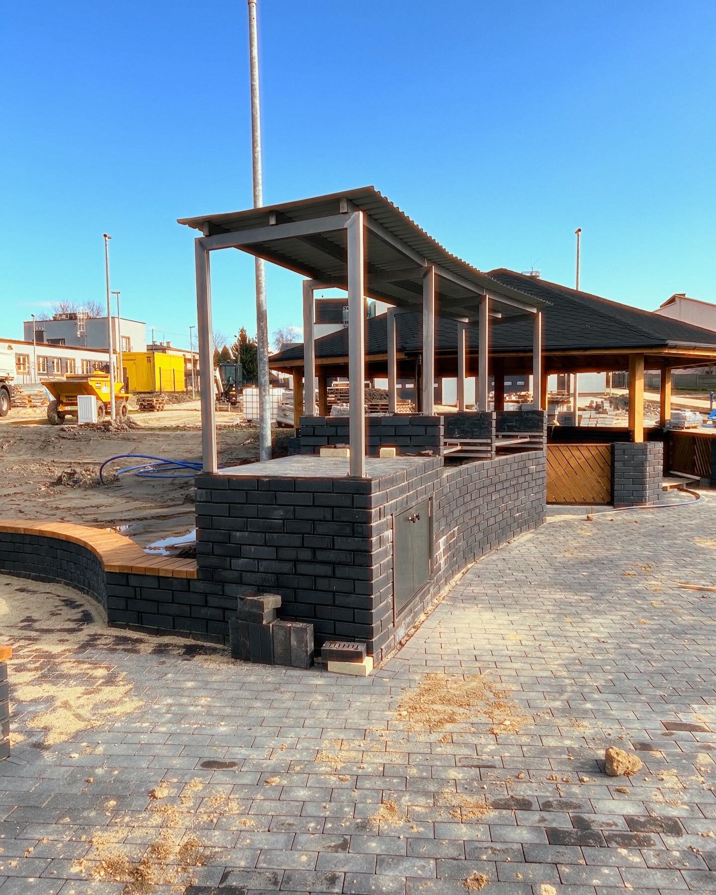 Wznoszona konstrukcja z ciemnej cegły z metalowym zadaszeniem, prawdopodobnie grill, widoczna na placu budowy z kostką brukową i żółtą maszyną budowlaną w tle.