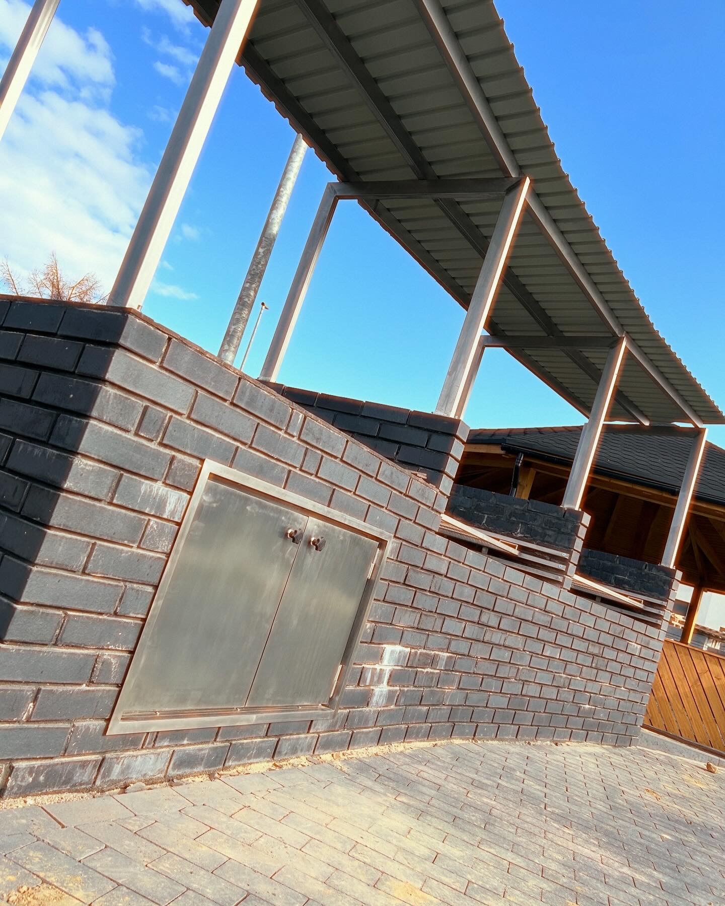 Murowana konstrukcja z szarej cegły z metalowymi drzwiczkami i zadaszeniem wspartym na stalowych słupach, widziana pod kątem z brukowej nawierzchni na tle błękitnego nieba.