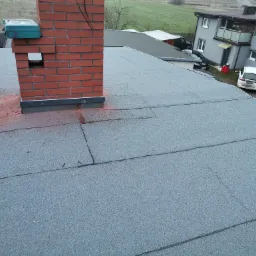 Szary dach pokryty papą z widocznym kominem z czerwonej cegły i narzędziem dekarskim obok, w tle zabudowa jednorodzinna i zieleń.