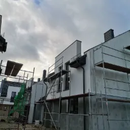 Budynek w trakcie ocieplania, widoczne rusztowania, częściowo otynkowana elewacja, zamontowane rynny. Na ziemi gruz i resztki materiałów budowlanych, pochmurne niebo.